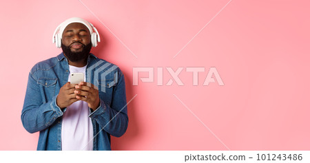 Delighted Black man enjoying awesome music, listening songs in headphones and holding smartphone, looking ecstatic, standing over pink background Delighted Black man enjoying awesome music, listening songs in headphones and holding smartphone, looking ecstatic, standing over pink background 101243486