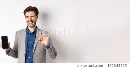 E-commerce and online shopping concept. Successful businessman making money on smartphone, showing empty phone screen and okay sign, winking happy at camera 101243525