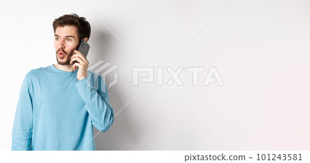 Technology concept. Intrigued young man looking left impressed, talking on mobile phone, calling someone, standing over white background Technology concept. Intrigued young man looking left impressed, talking on mobile phone, calling someone, standing over white background 101243581