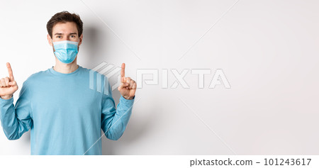 Coronavirus, health and quarantine concept. Handsome caucasian male model showing banner on top, pointing fingers up and wear medical mask, white background 101243617