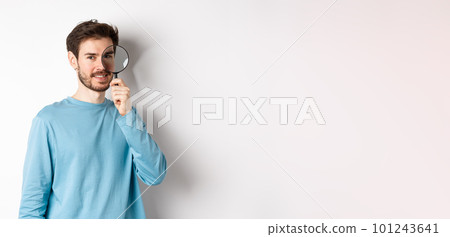 Smiling young man detective looking through magnifying glass, searching for promo offer, standing on white background Smiling young man detective looking through magnifying glass, searching for promo offer, standing on white background 101243641