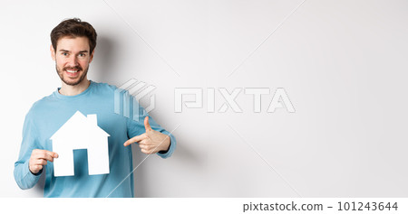 Real estate and insurance concept. Smiling young man pointing at paper house cutout and looking happy, buy apartment, standing over white background Real estate and insurance concept. Smiling young man pointing at paper house cutout and looking happy, buy apartment, standing over white background 101243644