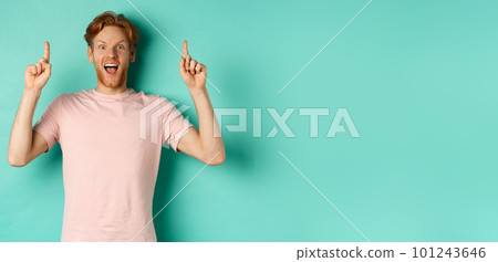 Cheerful redhead man in t-shirt pointing fingers up, staring in awe at camera and showing advertisement, standing over turquoise background Cheerful redhead man in t-shirt pointing fingers up, staring in awe at camera and showing advertisement, standing over turquoise background 101243646