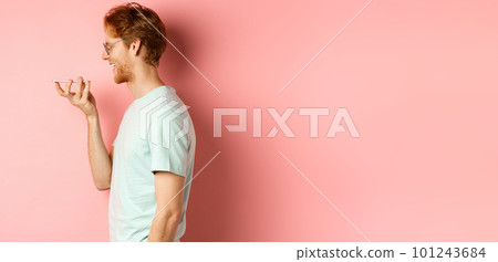 Profile portrait young man with red hair, smiling pleased while record voice message on smartphone, talking to virtual assistant, standing over pink background Profile portrait young man with red hair, smiling pleased while record voice message on smartphone, talking to virtual assistant, standing over pink background 101243684