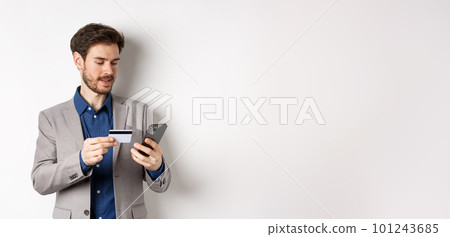 Online shopping. Smiling businessman paying with credit card on smartphone, sending money, standing in suit on white background 101243685