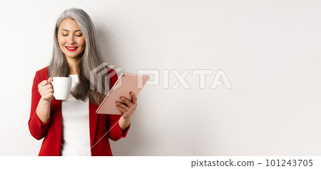 Business people concept. Asian mature businesswoman holding digital tablet and looking at coffee cup with pleased smile, standing over white background Business people concept. Asian mature businesswoman holding digital tablet and looking at coffee cup with pleased smile, standing over white background 101243705