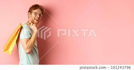 Carefree young man with red hair and glasses, walking with shopping bag over his shoulder and smiling, shopper buying presents, standing satisfied over pink background 101243796