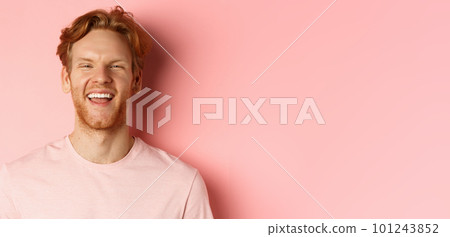 Close up of cheerful young man with messy red hair and beard, laughing and smiling, showing white teeth, looking happy at camera, standing over pink background 101243852