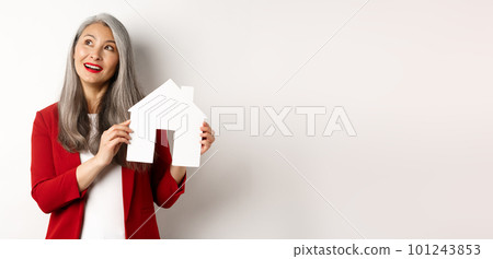 Dreamy senior woman thinking of buying property, showing paper house cutout and looking at upper left corner, standing over white background 101243853