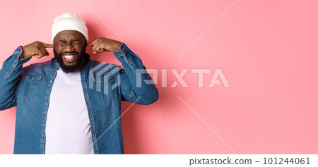 Displeased african-american man shut ears and grimacing from loud awful noise, disturb by annoying sound, standing over pink background 101244061