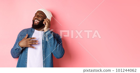 Happy african-american man talking on mobile phone, laughing and smiling, standing in beanie and denim shirt over pink background Happy african-american man talking on mobile phone, laughing and smiling, standing in beanie and denim shirt over pink background 101244062