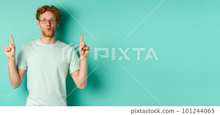 Impressed redhead man in glasses and t-shirt, checking out promo offer, pointing fingers up at copy space, staring at camera amazed, standing over turquoise background 101244065