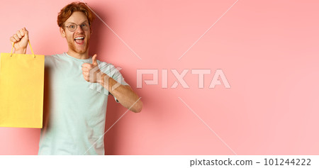 Cheerful redhead man in t-shirt and glasses pointing finger at shopping bag, showing shop with discounts, standing over pink background 101244222