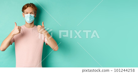 Covid-19, pandemic and lifestyle concept. Cheerful redhead guy in medical mask showing thumbs up in approval, like and praise product, standing over turquoise background Covid-19, pandemic and lifestyle concept. Cheerful redhead guy in medical mask showing thumbs up in approval, like and praise product, standing over turquoise background 101244258