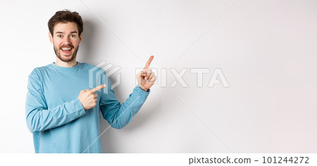 Cheerful smiling man pointing fingers right and showing banner or logo on white space for advertisements, recommending product, standing over studio background 101244272