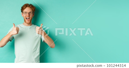 Excited young man with red hair, wearing glasses, showing thumbs-up and agree or praise something, smiling amazed and saying yes, standing over turquoise background 101244314