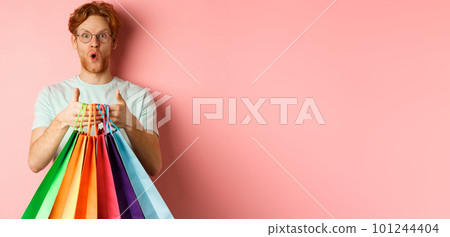 Young handsome man amazed with special discounts, buying gifts, holding shopping bags and standing over pink background Young handsome man amazed with special discounts, buying gifts, holding shopping bags and standing over pink background 101244404