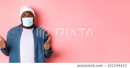 Coronavirus, lifestyle and social distancing concept. Black hipster guy in beanie and medical mask showing thumbs-up, praising product, looking left and smiling, pink background Coronavirus, lifestyle and social distancing concept. Black hipster guy in beanie and medical mask showing thumbs-up, praising product, looking left and smiling, pink background 101244431