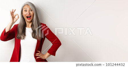 Happy asian mature woman in red blazer showing okay sign, smiling amazed, praising something awesome, standing over white background 101244432