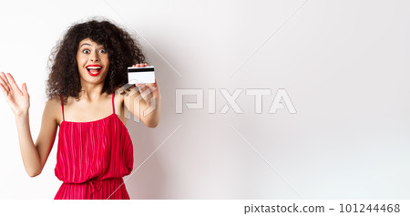 Shopping. Amazed curly-haired woman in red dress, showing plastic credit card and scream from excitement, standing on white background 101244468
