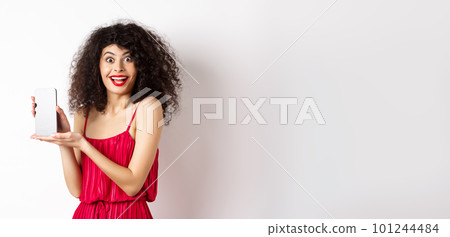Fashionable lady in red dress and makeup, showing mobile phone screen and smiling, introduce smartphone application, standing over white background 101244484