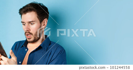 Close-up of man looking shocked and reading message on smartphone, gasping and stare startled at screen, standing on blue background 101244558