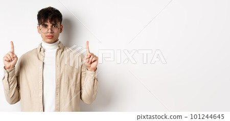 Handsome hipster guy in glasses pointing fingers up at empty space, looking serious at camera, standing on white background Handsome hipster guy in glasses pointing fingers up at empty space, looking serious at camera, standing on white background 101244645