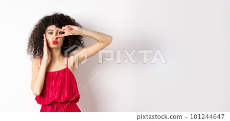 Beauty and fashion. Stylish curly-haired woman in red dress showing v-sign on eye and pucker lips, kissing you, standing on white background 101244647