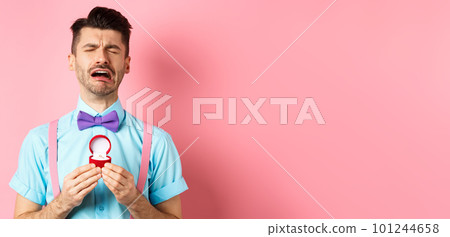 Valentines day. Heartbroken guy crying and holding engagement ring, sobbing from break-up, standing over pink background 101244658