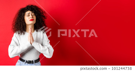 Desperate young woman pleading God, making wish and supplicating, holding hands in pray with eyes close and begging face, standing on red background 101244738