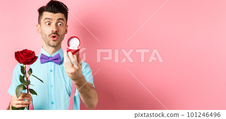 Valentines day. Romantic funny guy going to make wedding proposal, asking to marry him, holding red rose and looking at engagement ring with excitement, pink background Valentines day. Romantic funny guy going to make wedding proposal, asking to marry him, holding red rose and looking at engagement ring with excitement, pink background 101246496