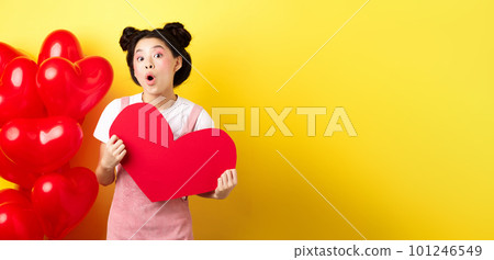 Happy Valentines day. Silly asian girl showing big red heart postcard and looking amazed, standing near red romantic balloons, yellow background 101246549