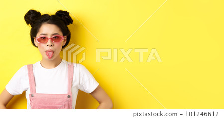 Summer and fashion concept. Silly asian teen girl showing tongue, wearing sunglasses, standing on yellow background Summer and fashion concept. Silly asian teen girl showing tongue, wearing sunglasses, standing on yellow background 101246612