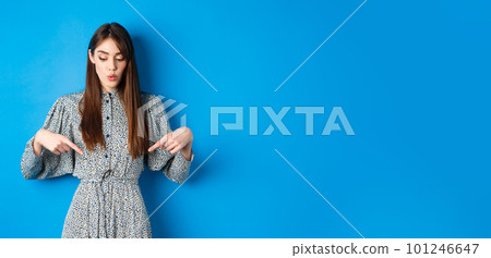 Intrigued beautiful girl in dress say wow, looking and pointing down at advertisement, showing promo with interested face, blue background 101246647