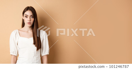 Shocked and startled young woman freeze in awe, open mouth and look at camera amazed, standing in dress on beige background 101246787