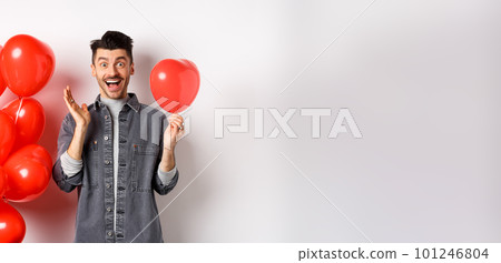 Valentines day concept. Cheerful young man celebrating love holiday, standing near red heart balloons and looking surprised, scream of joy, white background Valentines day concept. Cheerful young man celebrating love holiday, standing near red heart balloons and looking surprised, scream of joy, white background 101246804