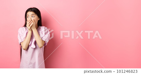 Shocked and startled asian woman looking aside at logo, covering mouth with hands speechless, standing in dress on pink background 101246823