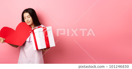 Valentines day concept. Romantic asian teen girl receive gifts on white day from lover, hugging big red heart card and present in box, smiling sensual, pink background Valentines day concept. Romantic asian teen girl receive gifts on white day from lover, hugging big red heart card and present in box, smiling sensual, pink background 101246830