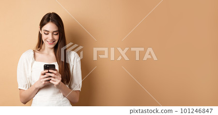 Young woman texting message on mobile phone, smiling and reading smartphone screen, standing on beige background Young woman texting message on mobile phone, smiling and reading smartphone screen, standing on beige background 101246847