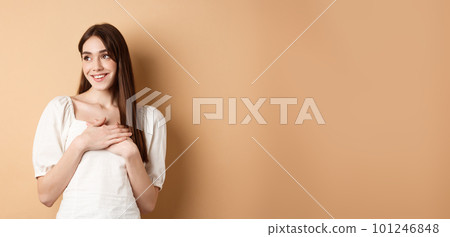 Romantic young girl in dress holding hands on heart, smiling and looking at empty space thankful, feeling gratitude, standing on beige background 101246848