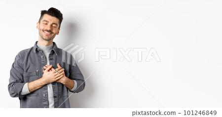Romantic happy man holding hands on heart and smiling with closed eyes, dreaming of something, feeling nostalgic from sweet memories, standing on white background 101246849