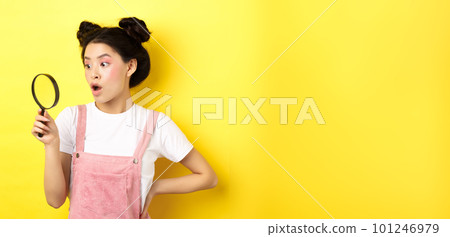 Excited asian woman with bright glamour makeup, look through magnifying glass amazed, staring aside, standing on yellow background 101246979