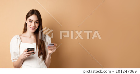 Smiling caucasian woman paying for online order, holding plastic credit card and mobile phone, looking carefree at camera, beige background 101247069
