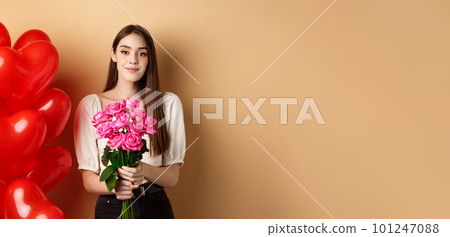 Beautiful girl holding bouquet of pink roses and smiling at camera, going on romantic date, standing near valentines heart balloons, beige background Beautiful girl holding bouquet of pink roses and smiling at camera, going on romantic date, standing near valentines heart balloons, beige background 101247088