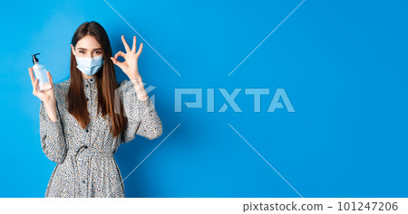 Covid-19, social distancing and healthcare concept. Beautiful girl in medical mask showing bottle antiseptic or hand sanitizer, make okay sign, recommend product, standing on blue background 101247206