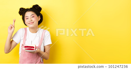 Holidays and celebration. Positive asian birthday girl cross fingers, making wish with b-day cake and lit candle, smiling happy at camera, yellow background 101247236