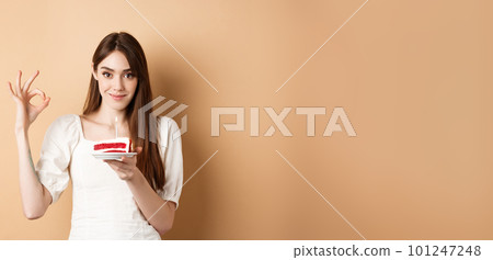 Pretty birthday girl holding cake and showing OK gesture, smiling pleased, enjoying bday celebration, beige background 101247248