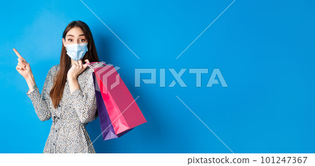 Covid-19, pandemic and lifestyle concept. Excited woman wears medical mask on shopping, pointing left corner logo, holding bags over shoulder, blue background 101247367