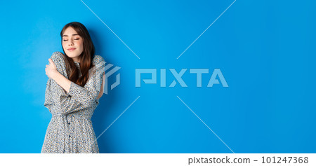 Romantic young natural woman in dress close eyes, smiling and hugging herself with love and tenderness, standing on blue background 101247368