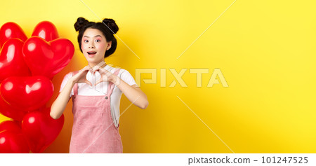 Happy asian girl showing heart sign near romantic balloons, waiting for true love on Valentines day, looking hopeful at camera, yellow background 101247525
